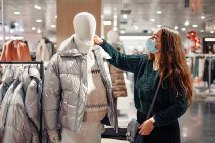 Young brunette lady in face mask chooses warm clothes during pandemic, buying puffy winter jacket